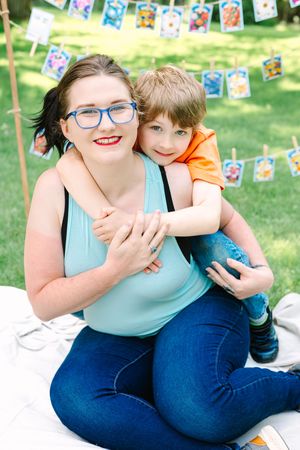 Two people share a warm embrace outdoors with colorful banners hanging in the background during a summer day.