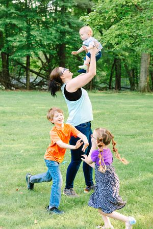 A joyful outdoor family moment with four people playing and laughing together on a green lawn surrounded by trees.