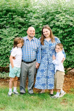 A family of four stands together outdoors against green foliage on a sunny day.