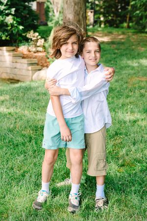 Two young children in casual summer clothes standing together and embracing on a grassy lawn with trees in background.