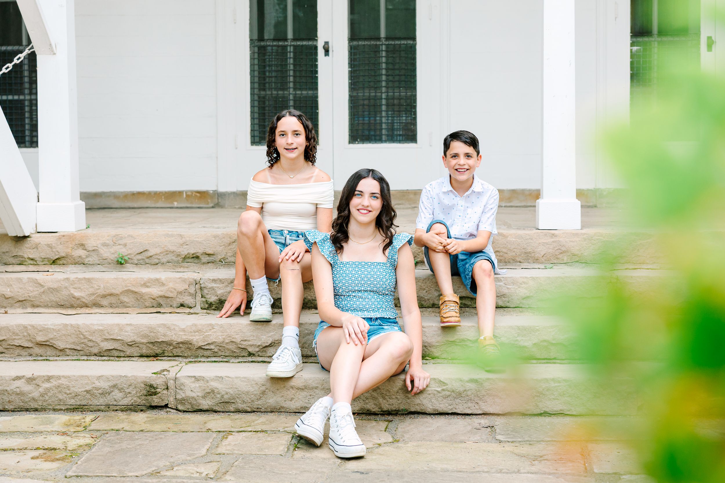 Family sitting and relaxing on rustic porch steps surrounded by lush green foliage on a sunny afternoon.