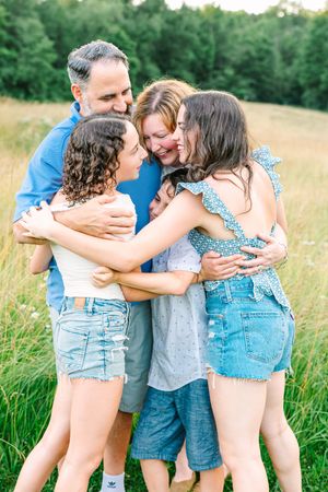 Family embraces in a warm group hug while standing in a sunlit summer meadow with green trees in background.