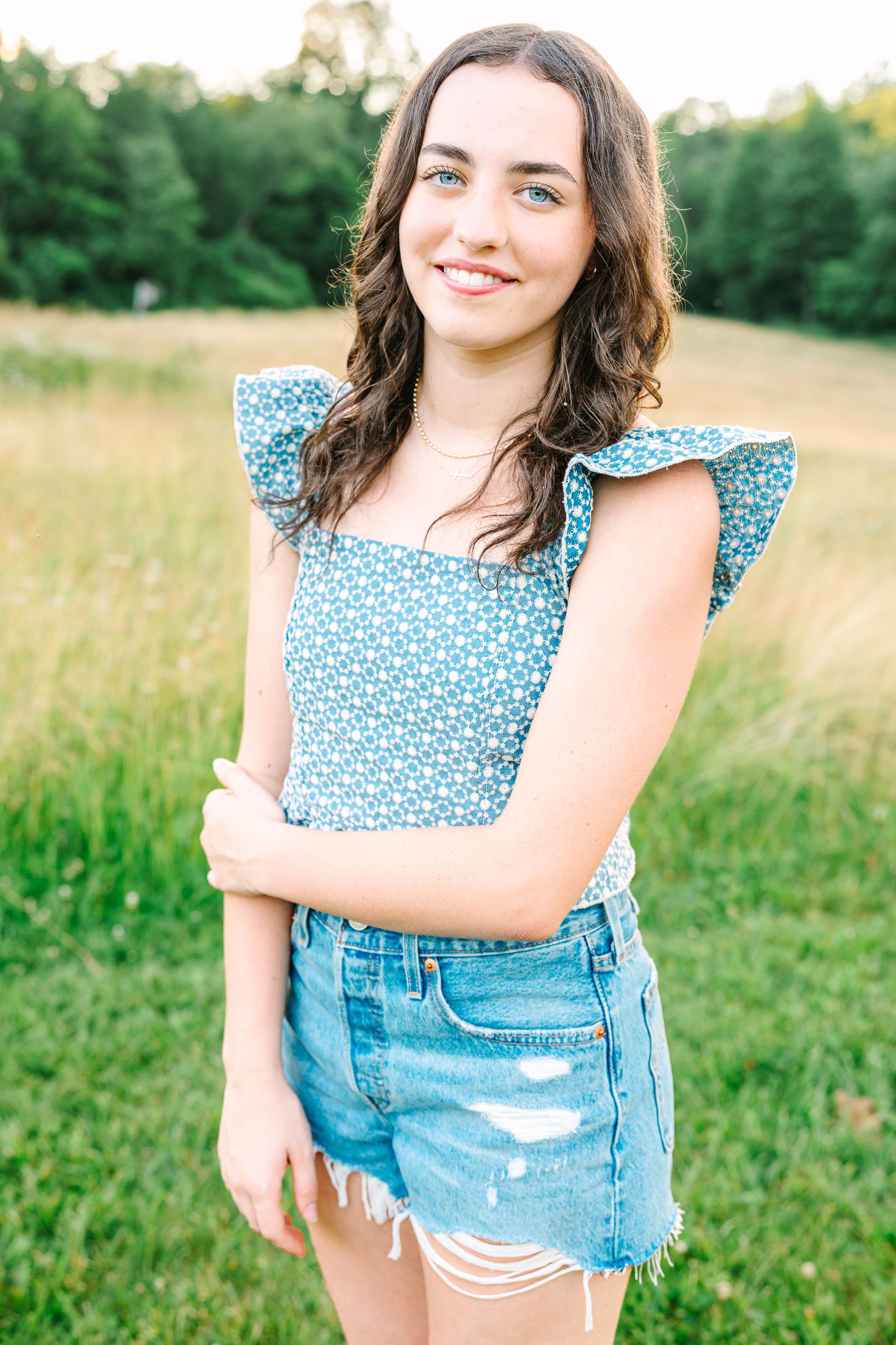 Young woman in denim shorts and floral top poses in a sunny meadow with flowing grass.
