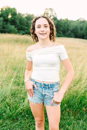 Young person stands in white top and jean shorts in a grassy summer field with trees behind them.