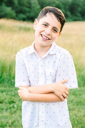Young boy in white polka dot shirt smiling and posing playfully in a summer field.