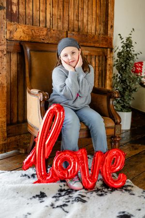 Grey-clad figure sits on wooden chair with large red LOVE balloon letters on white carpet near potted plant.