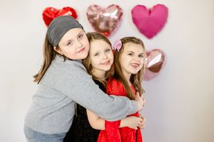 Two people wearing red and gray outfits embrace and smile at the camera in front of heart-shaped decorations.
