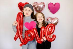 Two people holding a large red metallic LOVE balloon with heart decorations against a neutral background.