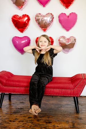 A child in a black outfit plays on a red chaise lounge with heart-shaped balloons decorating the white wall behind them.