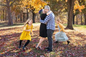 A couple in autumn attire playfully throws colorful fall leaves in a sunlit wooded park setting during a photo session.