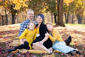 A family enjoys a warm autumn day while posing together among fallen yellow leaves in a park setting.