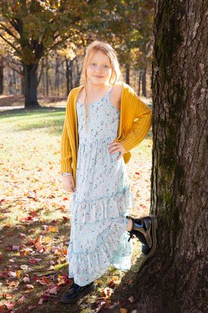 A light blue floral dress catches sunlight in a wooded autumn setting with fallen leaves scattered on the ground.