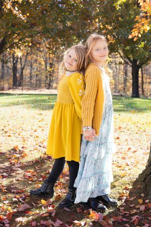 Two children in bright yellow and pale blue dresses stand amid falling autumn leaves in a sun-dappled park setting.