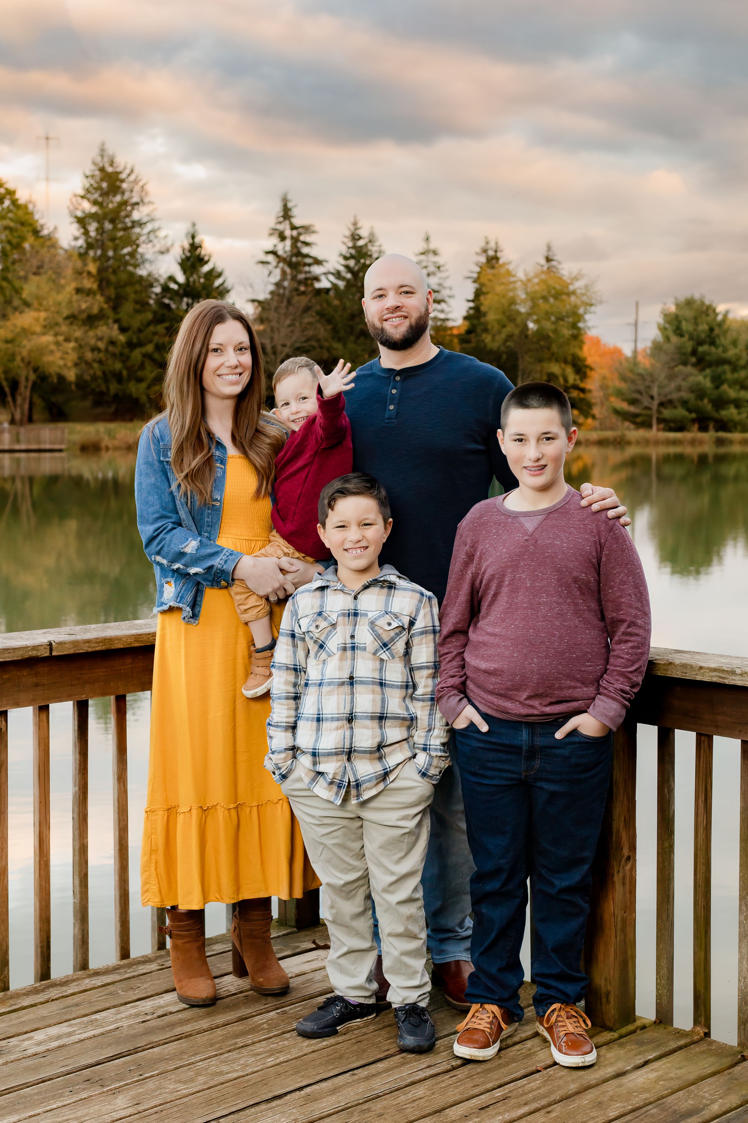A family dressed in yellow, burgundy and plaid poses for portraits on a wooden dock by a tranquil lake.