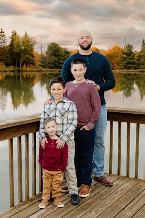 A fall family portrait standing on a wooden dock overlooking a lake with autumn trees in the background.