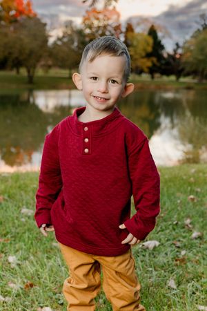A child wearing a burgundy sweater and mustard pants poses outdoors near a lake during autumn.