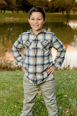Young person in plaid button-down shirt and khaki pants poses confidently by a pond on grassy lawn.