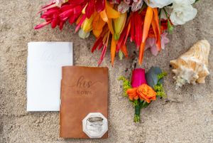 Wedding rings and vow books arranged with tropical flowers and seashells on sandy beach for destination ceremony.
