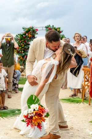 Romantic wedding kiss under a tropical floral arch with palm leaves and colorful bouquet on a sunny beach.