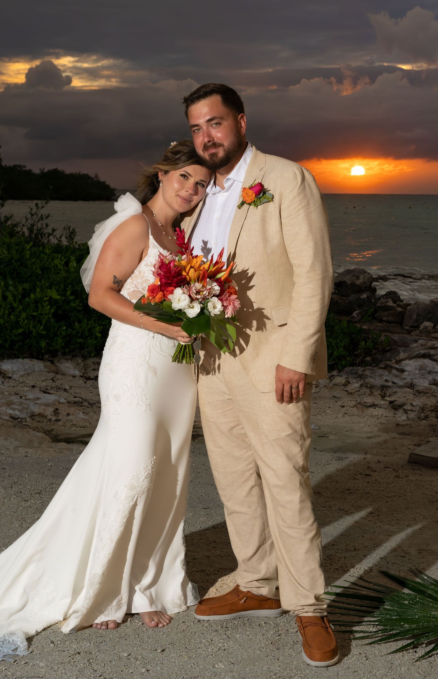 A couple poses together at sunset on a beach, the bride holding a vibrant red and white bouquet.