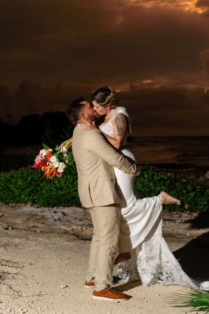 Couple shares romantic embrace on tropical beach during sunset with dramatic orange sky and colorful bouquet.