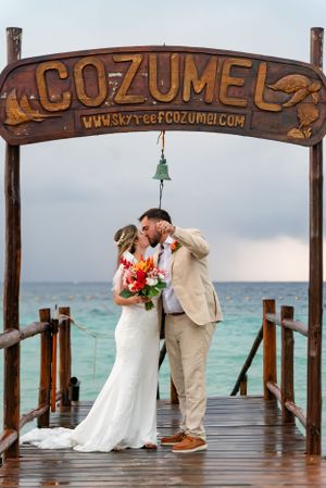 A wooden pier entrance with a Cozumel sign over a couple on their wedding day with turquoise ocean waters in background.