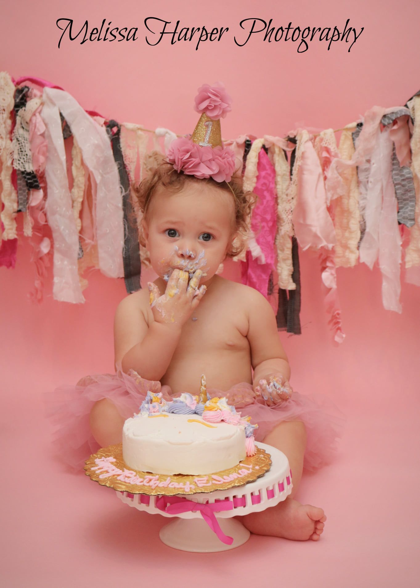 Cake smash photo with white cake on pink stand and fabric banner backdrop.