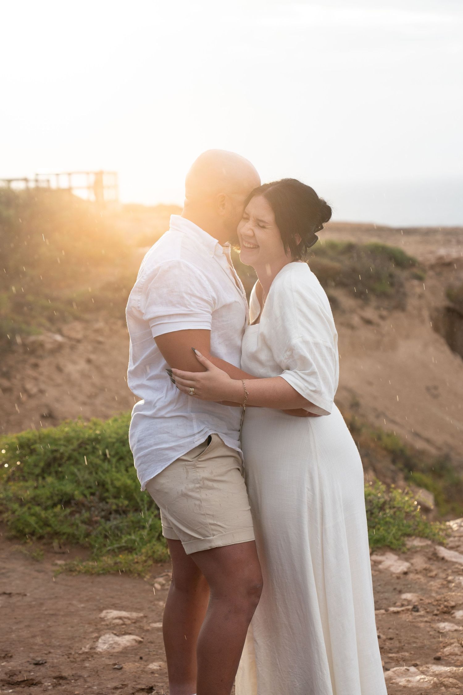 Loving couple embraces on a coastal cliff during golden hour sunset in matching white outfits.