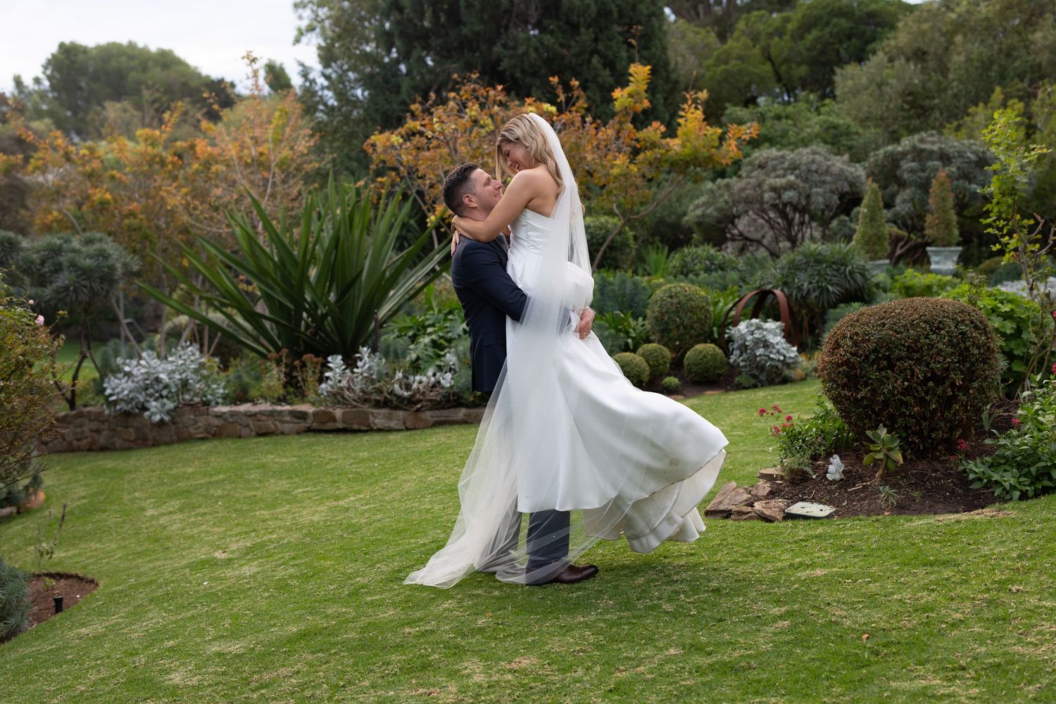 Romantic wedding couple twirls together on a lush garden lawn surrounded by scenic autumn foliage.