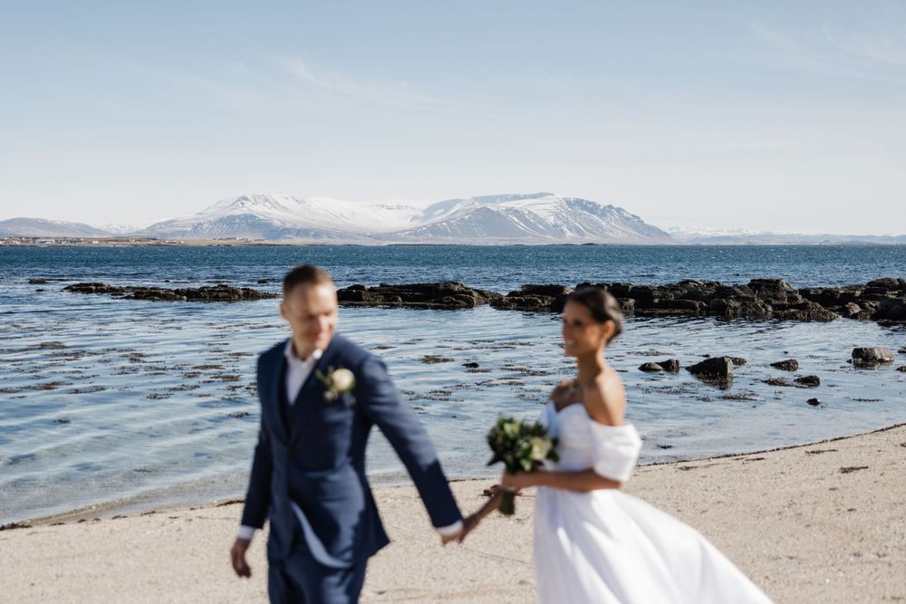 A couple walks along a scenic beach with snow-capped mountains in the background.