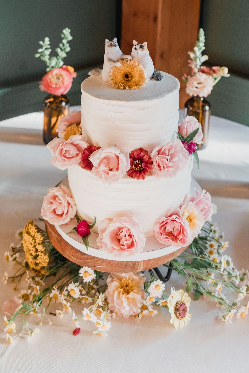Two-tier wedding cake decorated with pink roses, daisies and topped with porcelain cat figurines in photo by NH wedding photographer