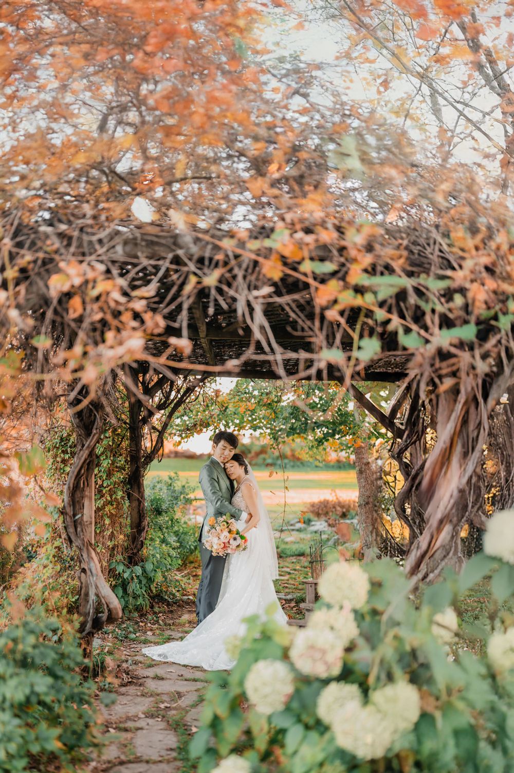 A couple embraces under a rustic wooden archway with autumn foliage and white hydrangeas in a garden setting at Trapp Lodge in Stowe, VT