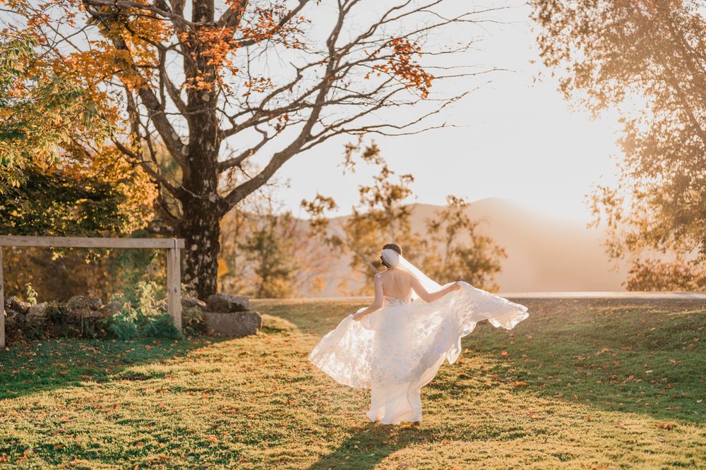 A flowing white dress twirls in golden sunlight beneath an autumn tree during sunset on a grassy hilltop by Maine wedding photographer