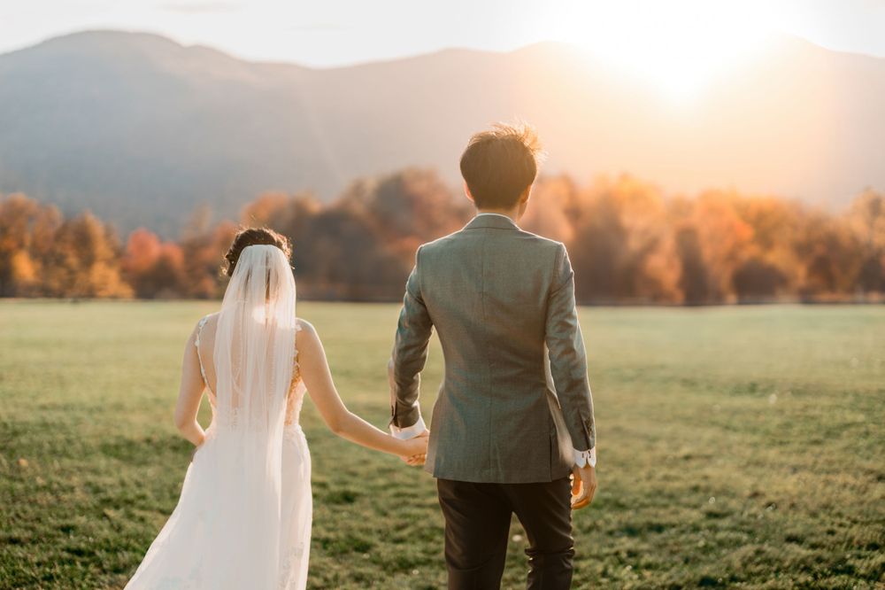 A couple in wedding attire holds hands looking at a sunset over mountains in an autumn meadow at Trapp Lodge by Stowe wedding photographer
