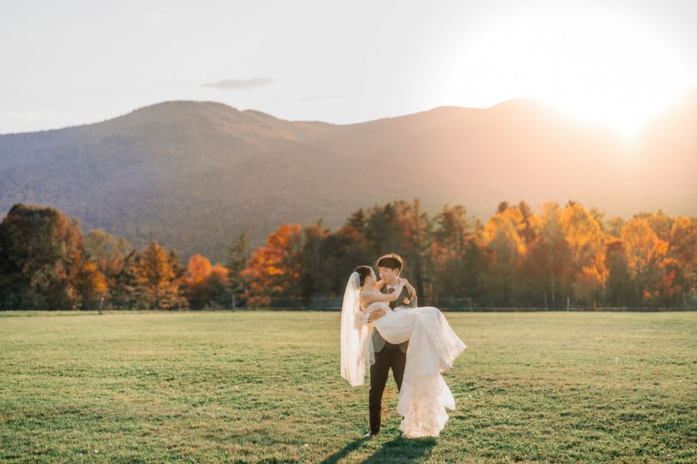 A couple stands in a meadow with autumn foliage and mountains at sunset in the background at Trapp Lodge in Stowe by VT wedding photographer
