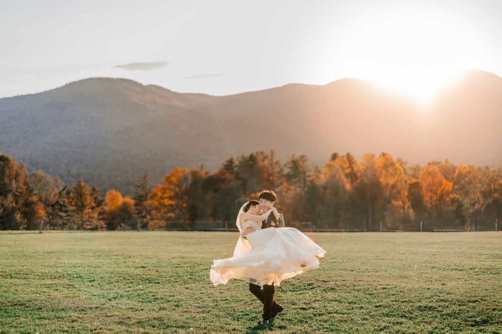 A bride in a flowing white dress twirls in a mountain meadow in the arms of the groom with autumn trees by Maine wedding photographer