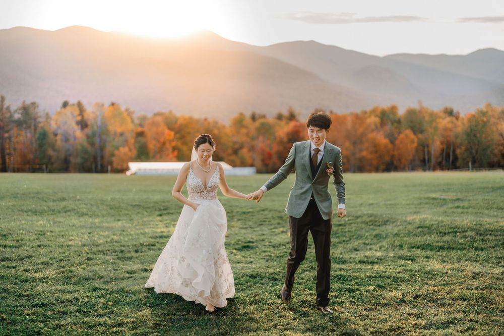 A couple holds hands running through a grassy field with autumn mountains and golden sunlight in in photo by Maine wedding photographer