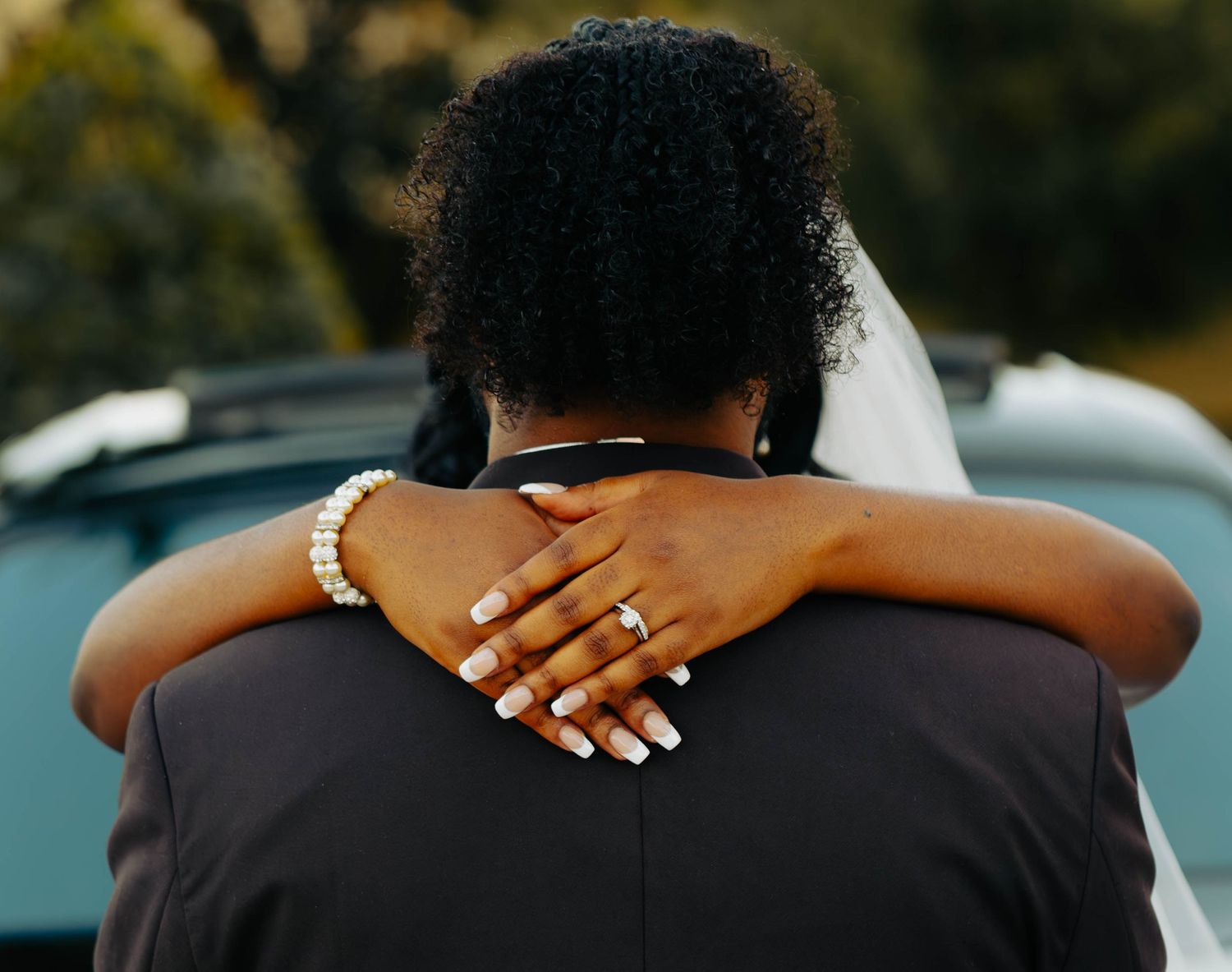 A bride and groom embrace from behind, showcasing a diamond ring and pearl bracelet outdoors.