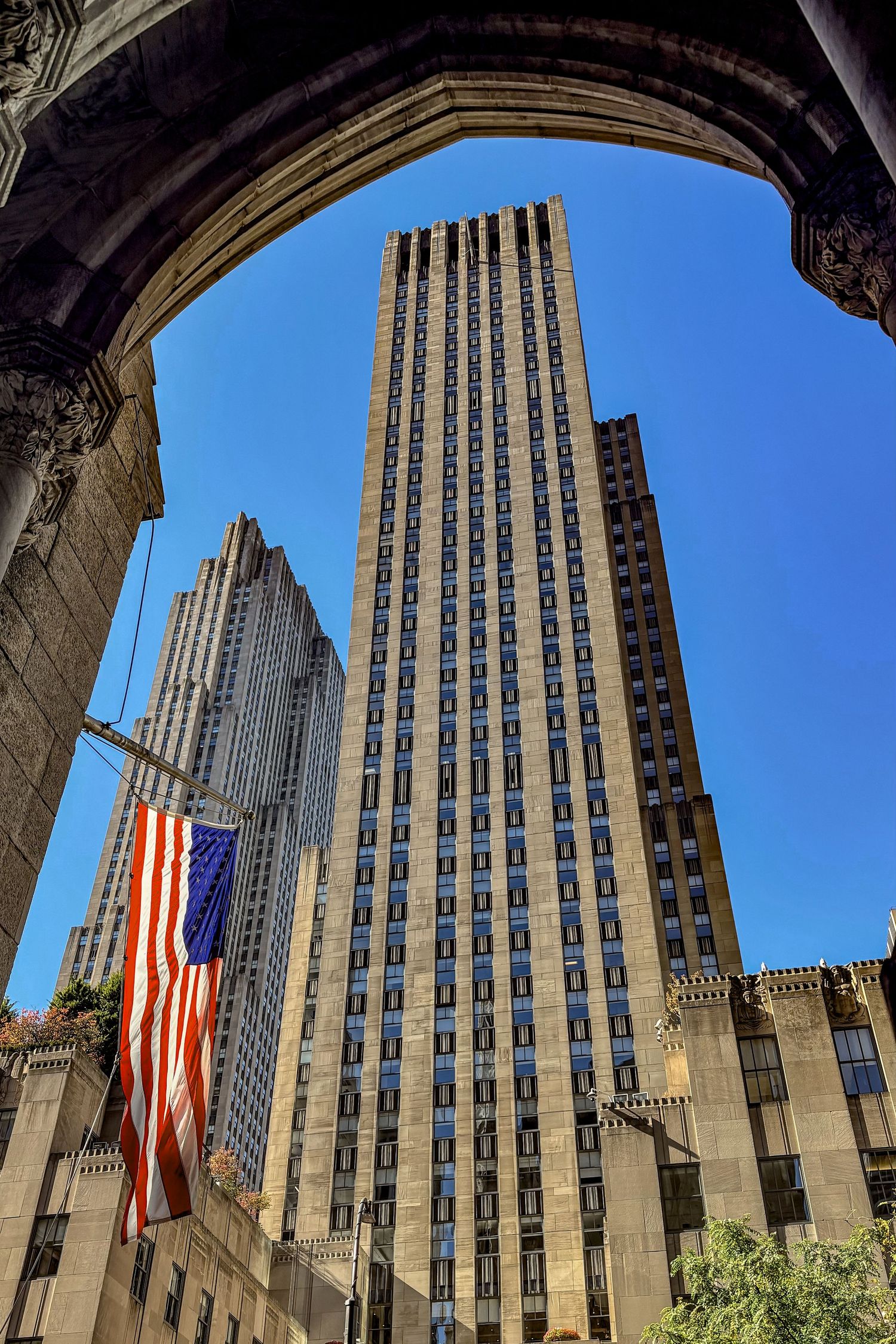 Looking up at towering skyscrapers through an archway with an American flag in New York City.