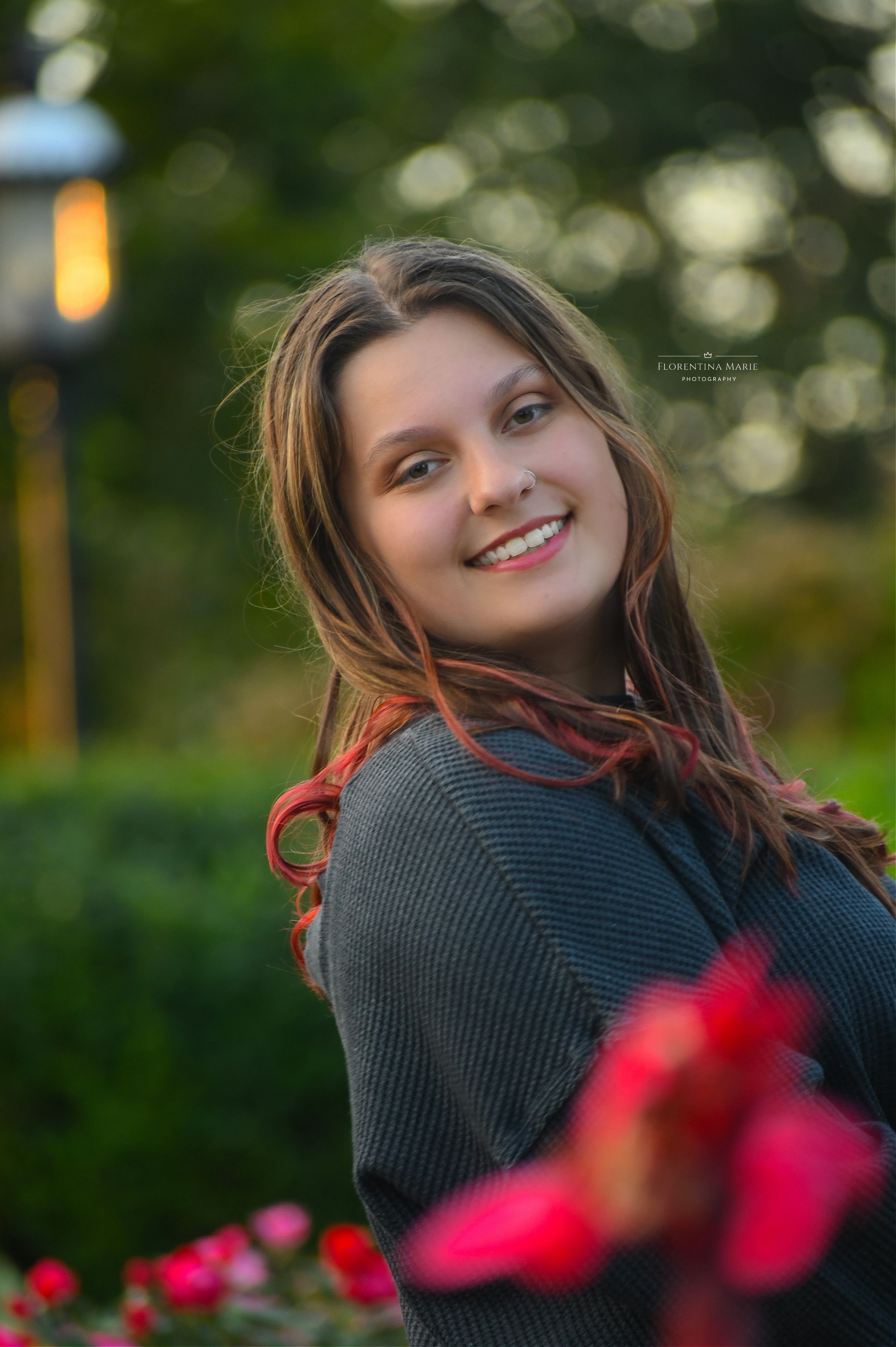 Portrait of someone with long brown hair smiling against a bokeh background with red flowers.