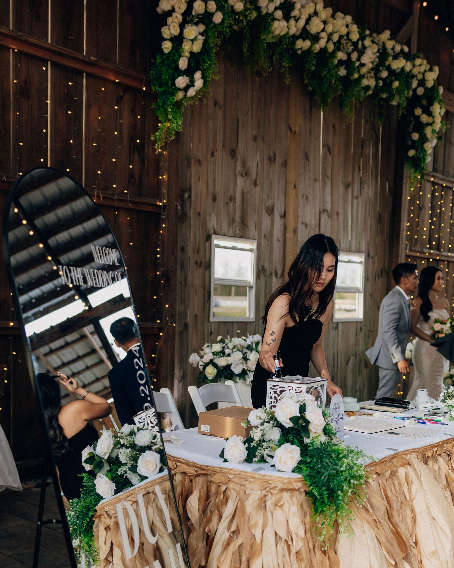 Wedding guest signs in at a decorated barn reception table with white floral arch and fairy lights overhead.