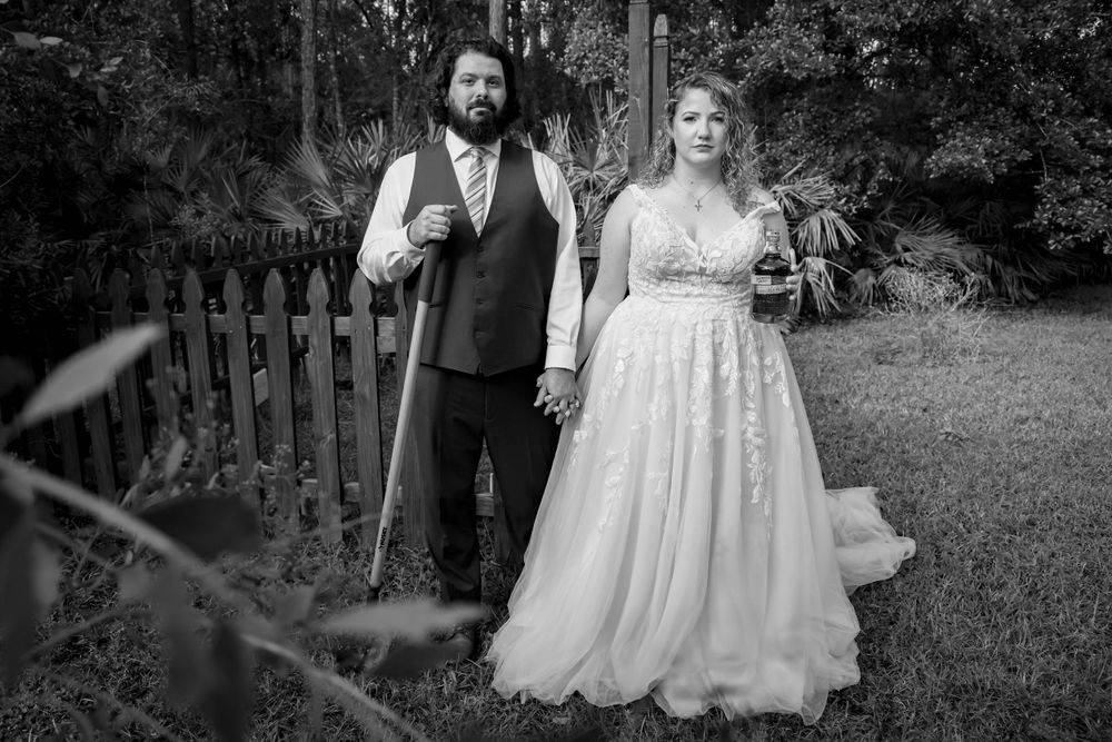 Black and white portrait of couple in wedding attire standing by rustic fence.