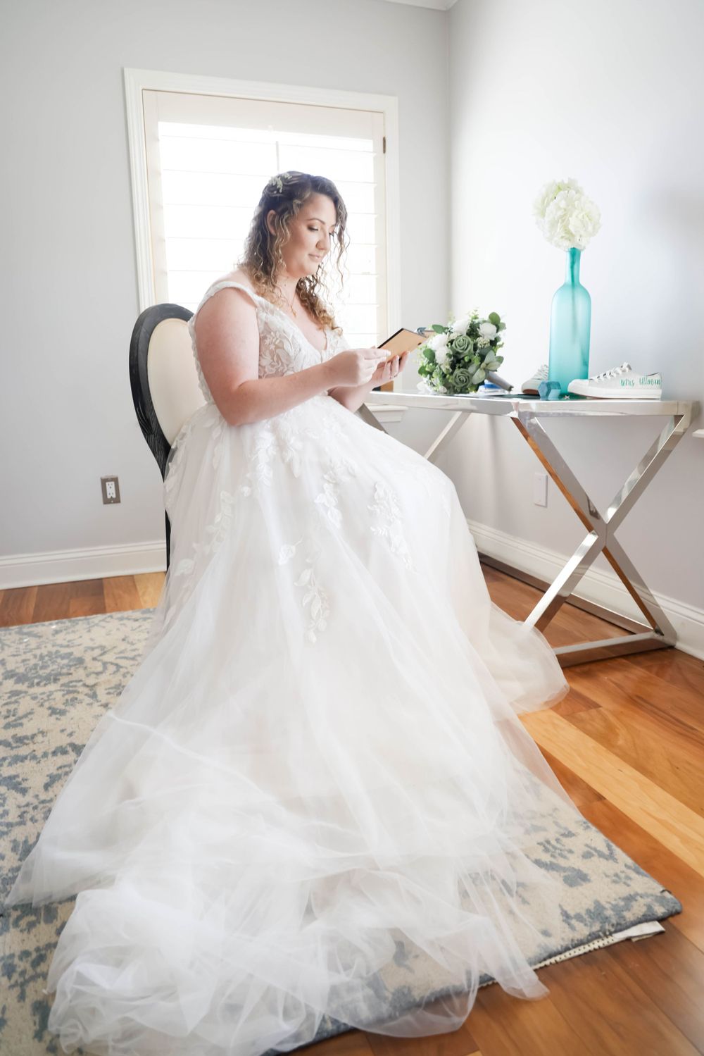 Person in elegant white wedding dress sitting at small table near window reading.