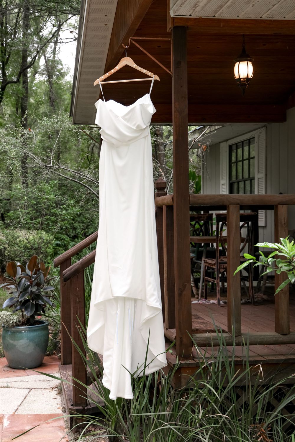 White wedding dress hanging on wooden porch with natural greenery and rustic decor.