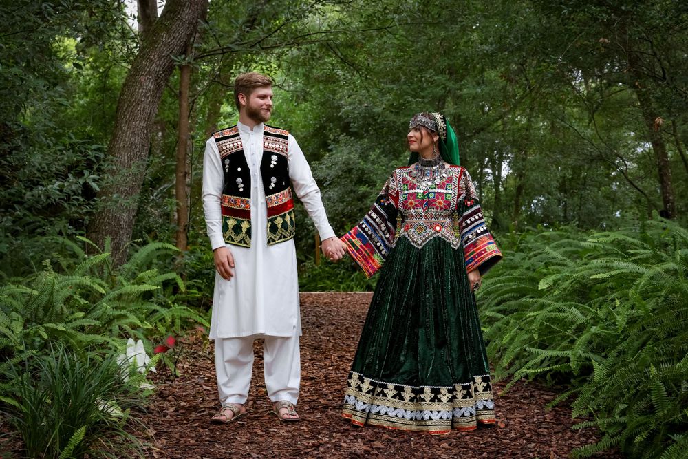 Couple in traditional Afghan wedding attire standing in lush garden setting.