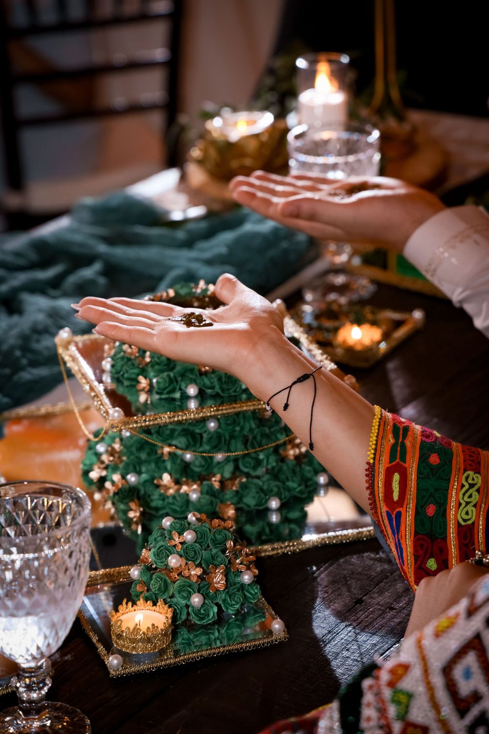 Hands reaching over a decorative green crystal tree with glowing candles and ornate table settings in warm lighting.