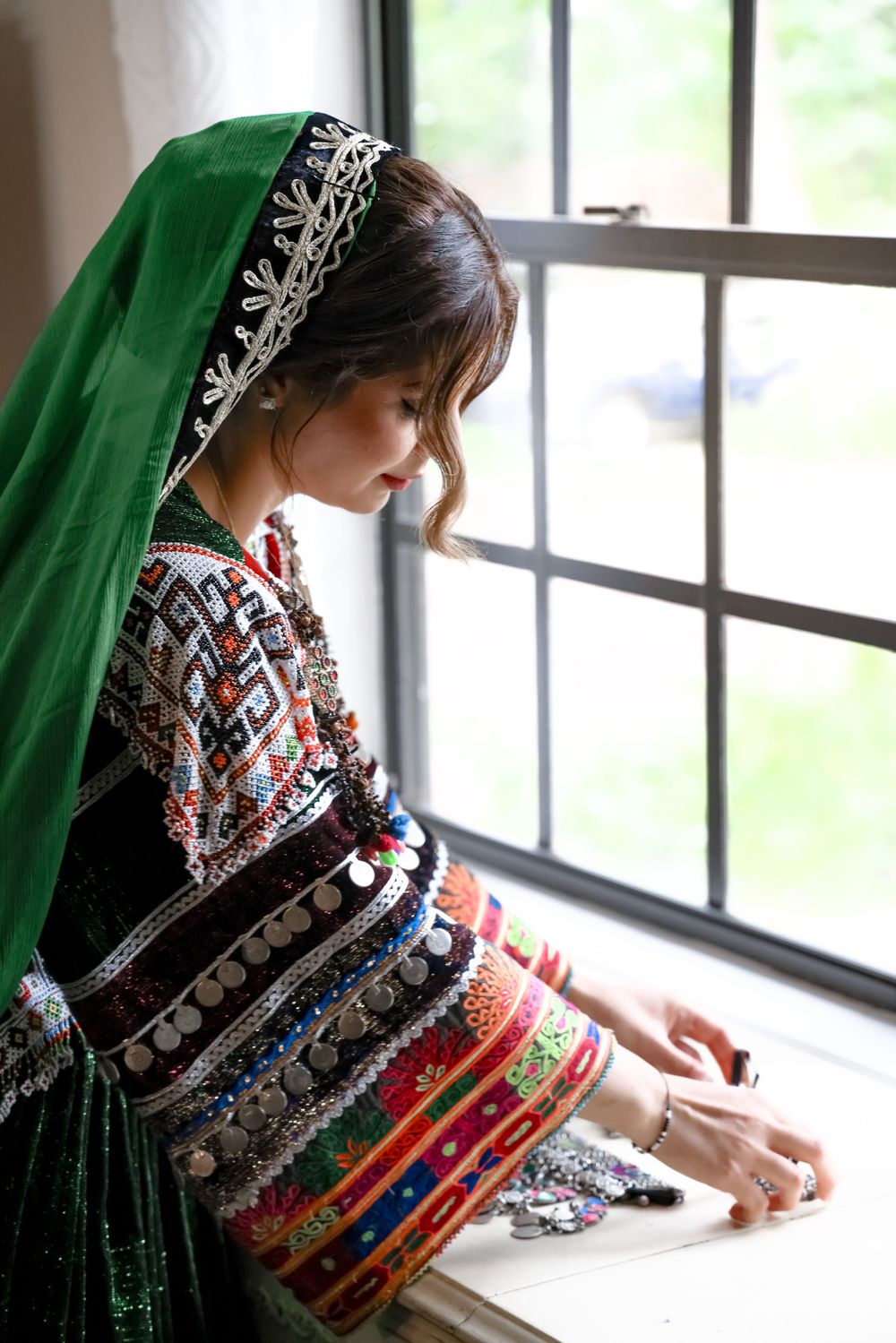 Traditional Afghan dress with intricate embroidery and green headscarf shown in window light.