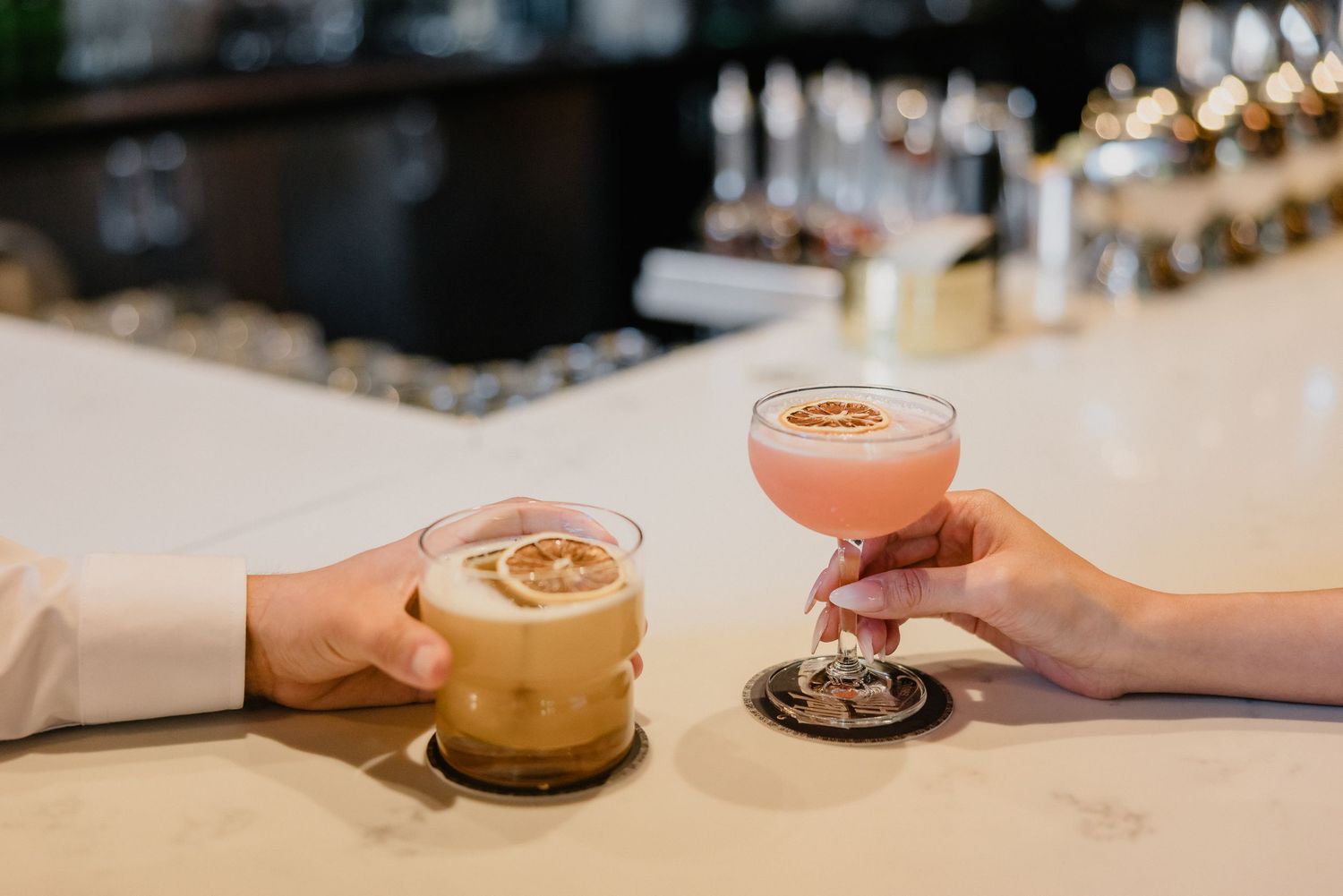 Two people toasting with cocktails at a contemporary bar counter, one drink appears creamy and one appears pink.