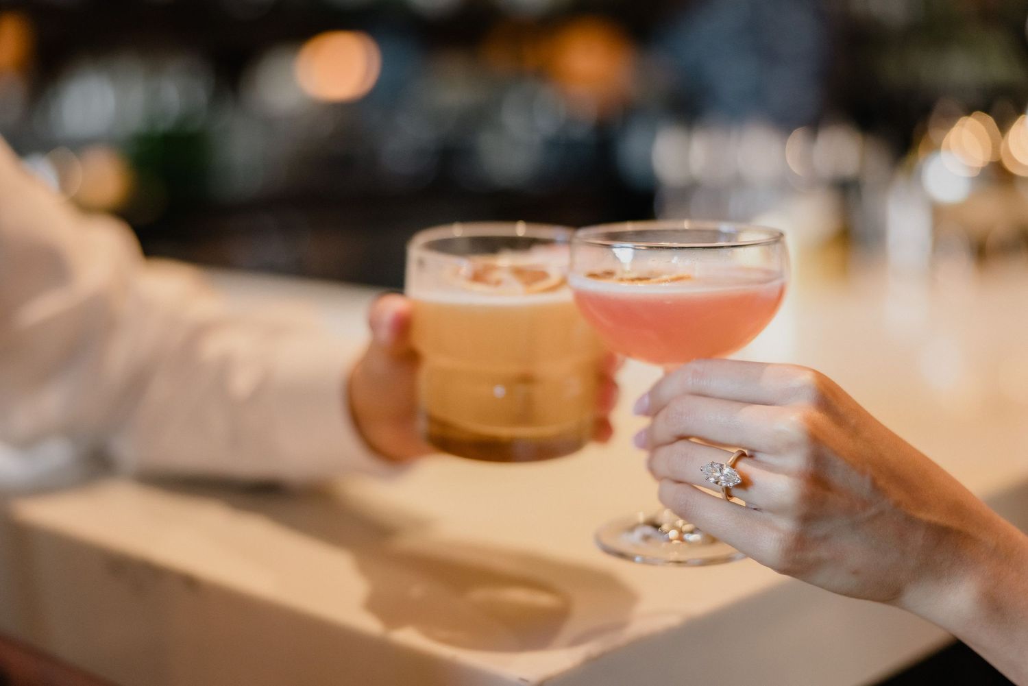 Hands holding up elegant cocktail glasses for a toast, featuring one creamy and one pink beverage at a bar.