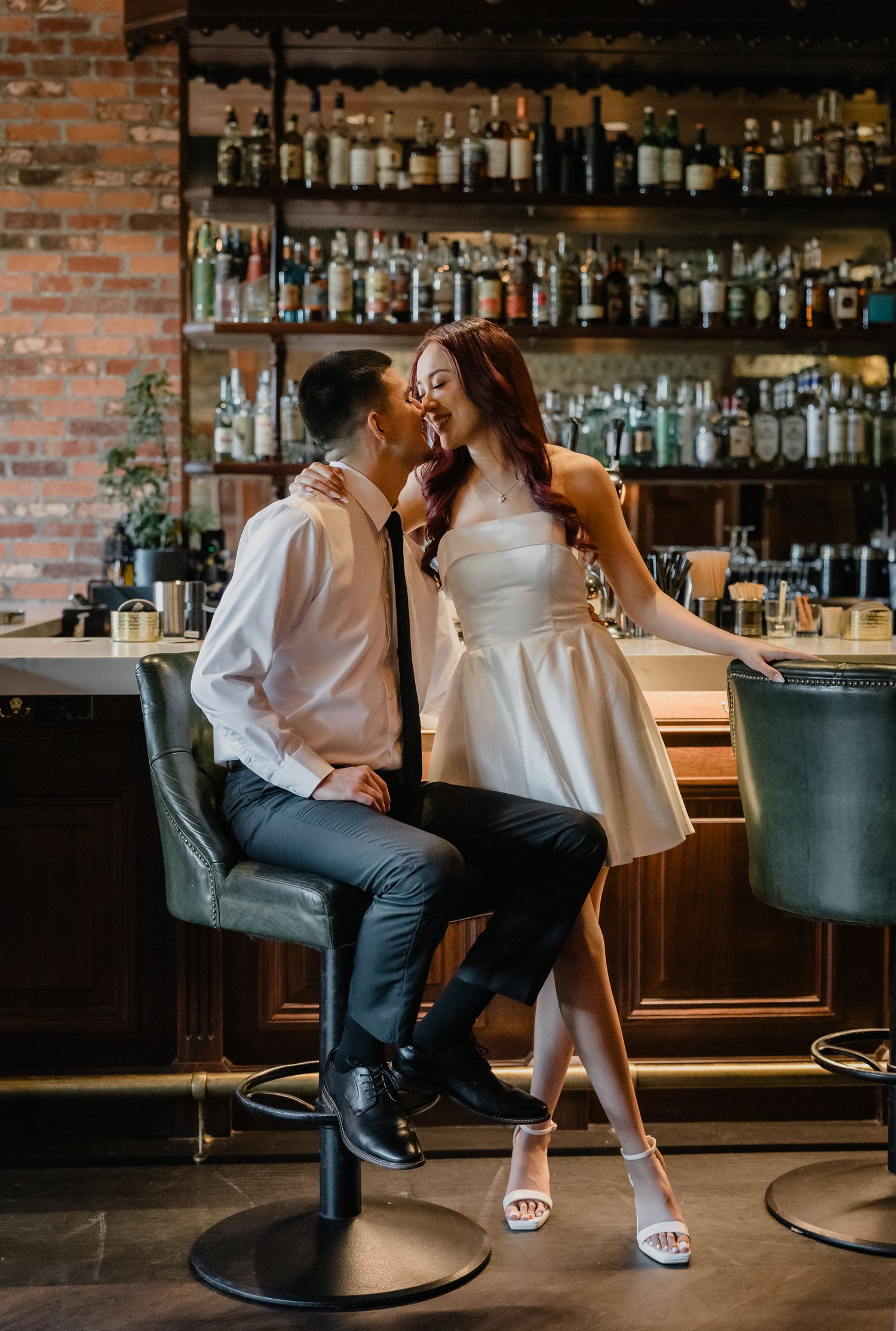 Romantic couple shares an intimate moment at an upscale bar with exposed brick walls and shelves of liquor bottles.